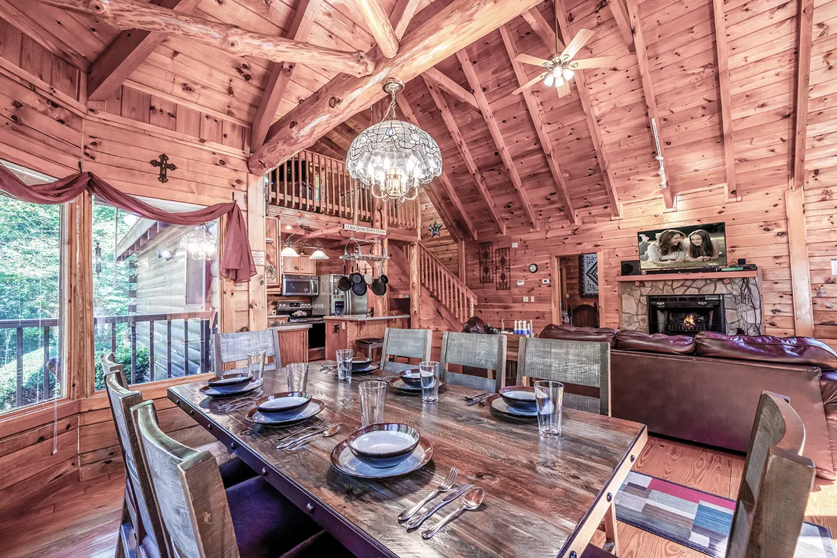 A view of a large wooden dining table with a chandelier above, a stone fireplace to the side, and an open kitchen area in the background. The scene radiates coziness and a space ideal for communal meals or game nights, with expansive windows that bring the outdoors in.