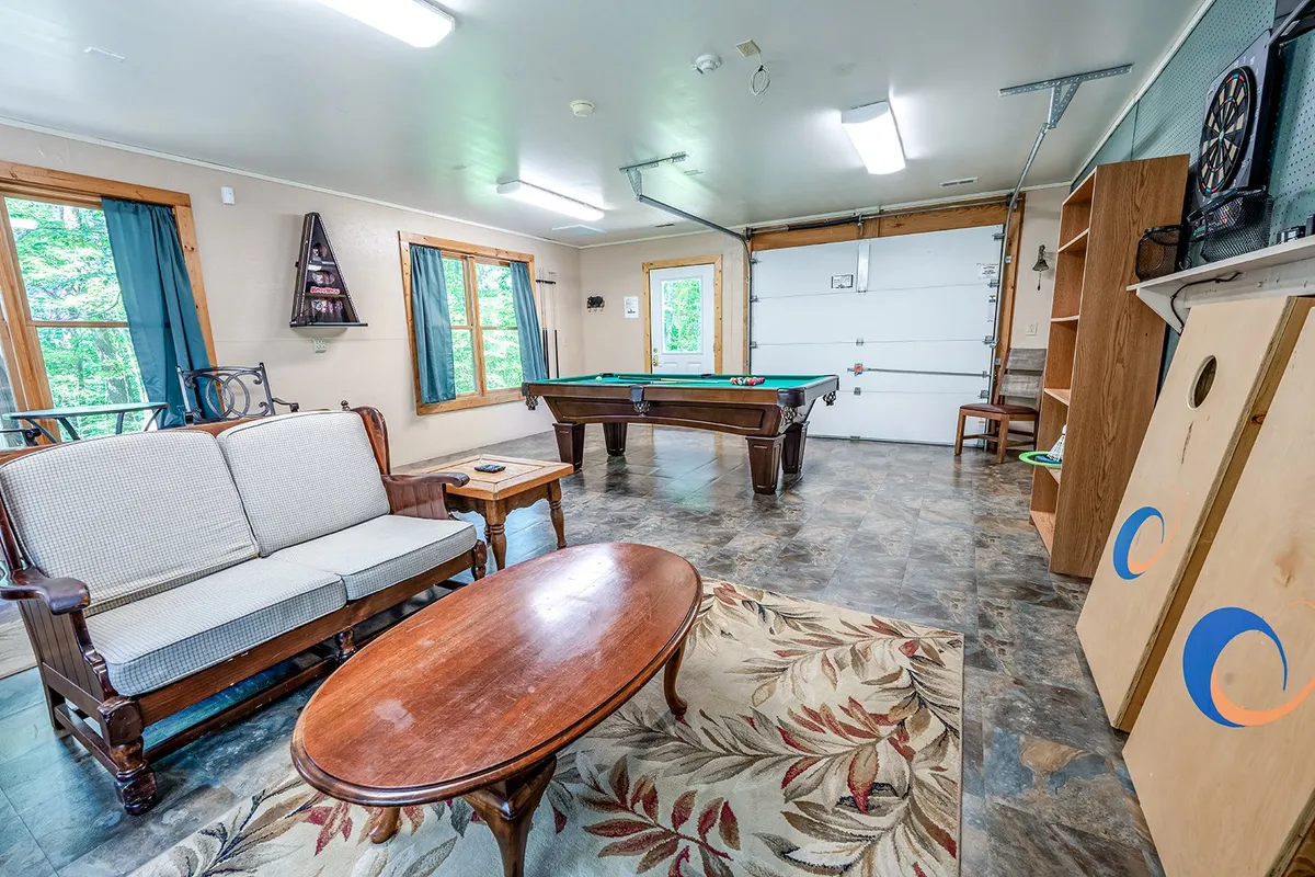 A casual and spacious game room with a pool table as the centerpiece, comfortable sofa and coffee table nearby, bright windows for natural light, and a dartboard on the wall. The arrangement invites both socializing and spirited gameplay.
