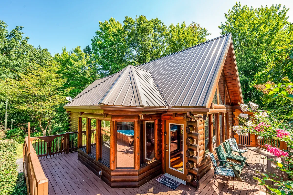 This exterior shot features the inviting log cabin structure of Steele Away, complete with a metal roof, deck seating, and lush greenery all around. The deck is visible as a welcoming space for families to gather after a day out enjoying local attractions.