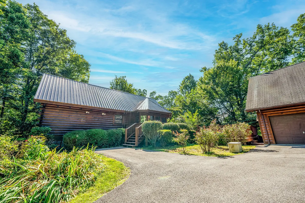 A welcoming log cabin set amidst lush greenery, with a driveway leading up to the home. The porch and surrounding trees evoke a sense of peaceful seclusion and easy access to nature, making it an ideal home base for outdoor adventures.