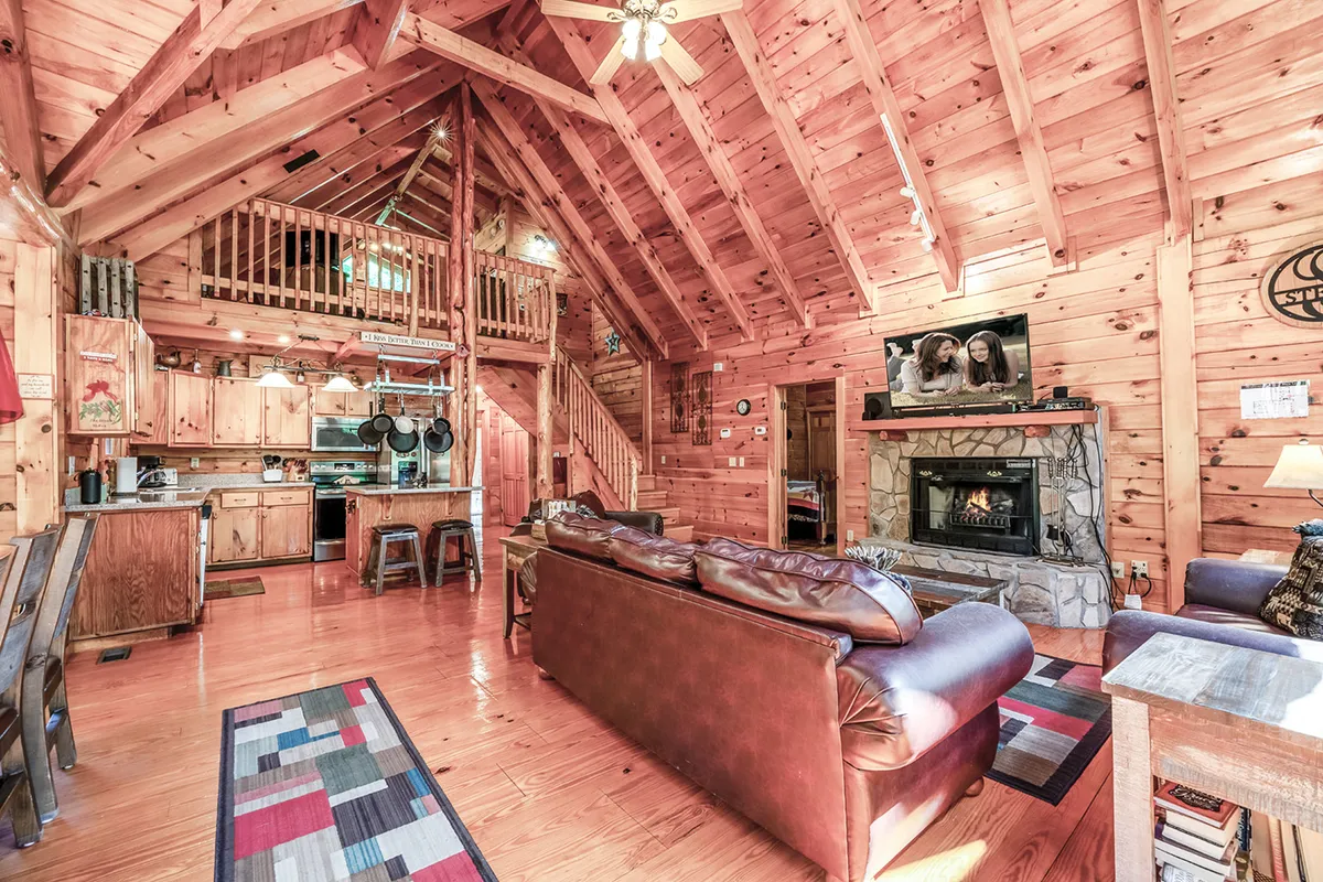 A spacious open-concept living area featuring a stone fireplace surrounded by comfortable seating, visible wooden beams, a fully equipped kitchen area, and large windows that fill the room with natural light. The arrangement encourages togetherness and relaxation, with easy flow between the living, dining, and kitchen spaces.