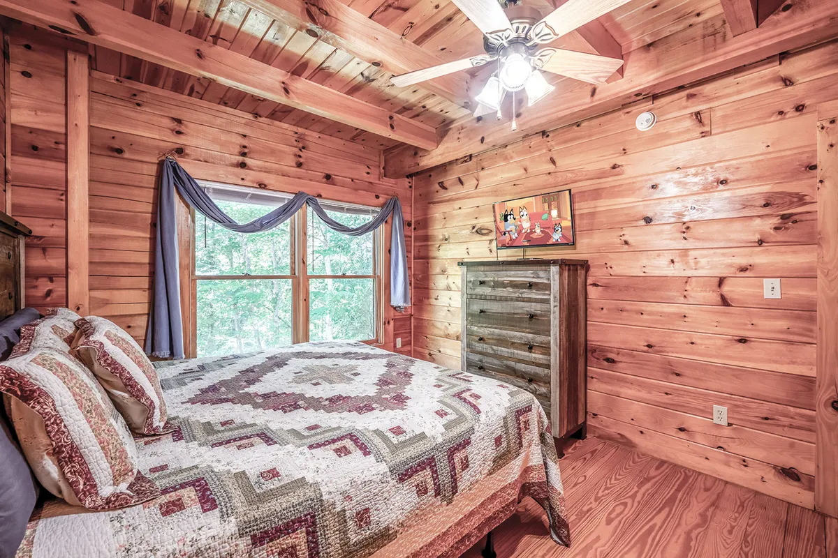 The photo highlights a king bedroom with a quilted bedspread, wooden furniture, large windows streaming in daylight, and a flat-screen TV mounted on the wall. The setting is cozy yet spacious, blending plush linens and comforting decor with modern conveniences.