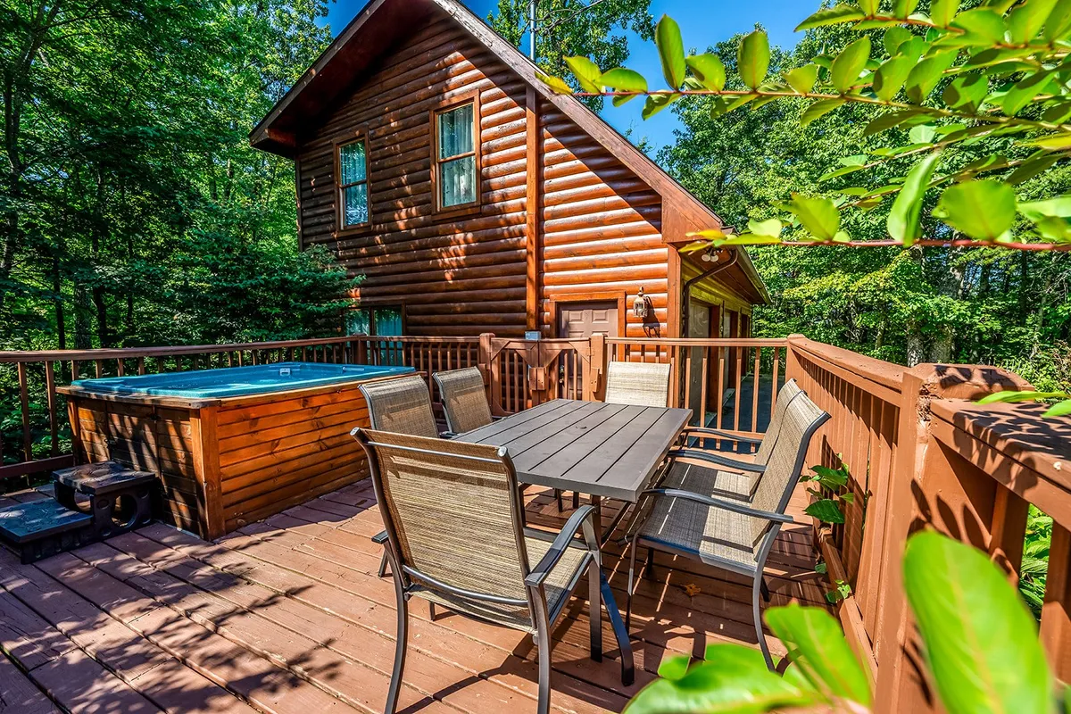 An exterior view of the spacious wooden deck, featuring a dining table and a bubbling hot tub, overlooking the scenic greenery. The log cabin and hot tub invite families to unwind together after busy days exploring Dollywood and nearby attractions.