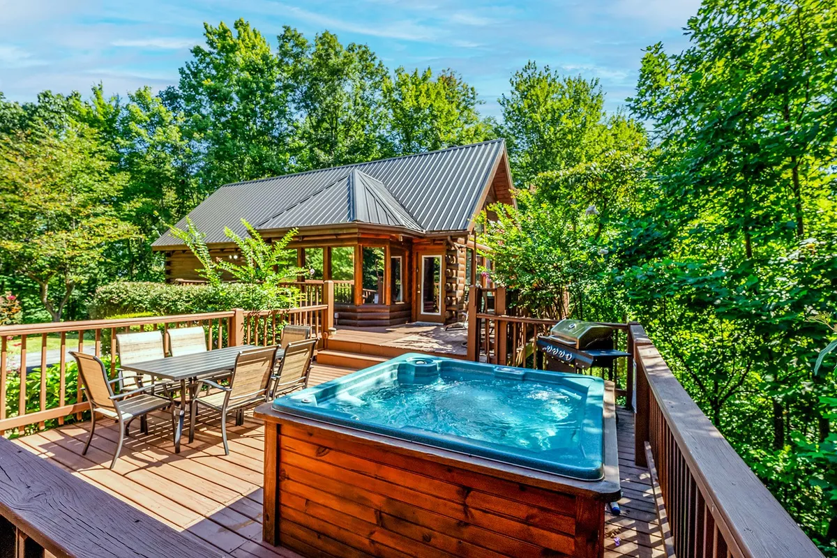 A spacious wooden deck nestled within lush greenery, featuring a private hot tub, a dining table set for six, and a grill. The area is surrounded by nature, offering a tranquil atmosphere perfect for relaxation or group gatherings.