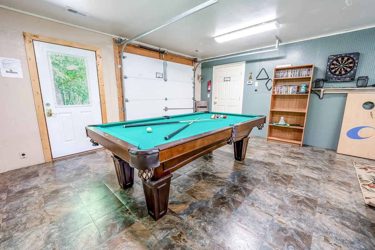 A garage-converted game room featuring a pool table in the foreground, a garage door to the side, organized shelves stocked with games and DVDs, and a cozy, relaxed setup ideal for entertainment. The space exudes a welcoming and playful vibe, with ample space for group activities.