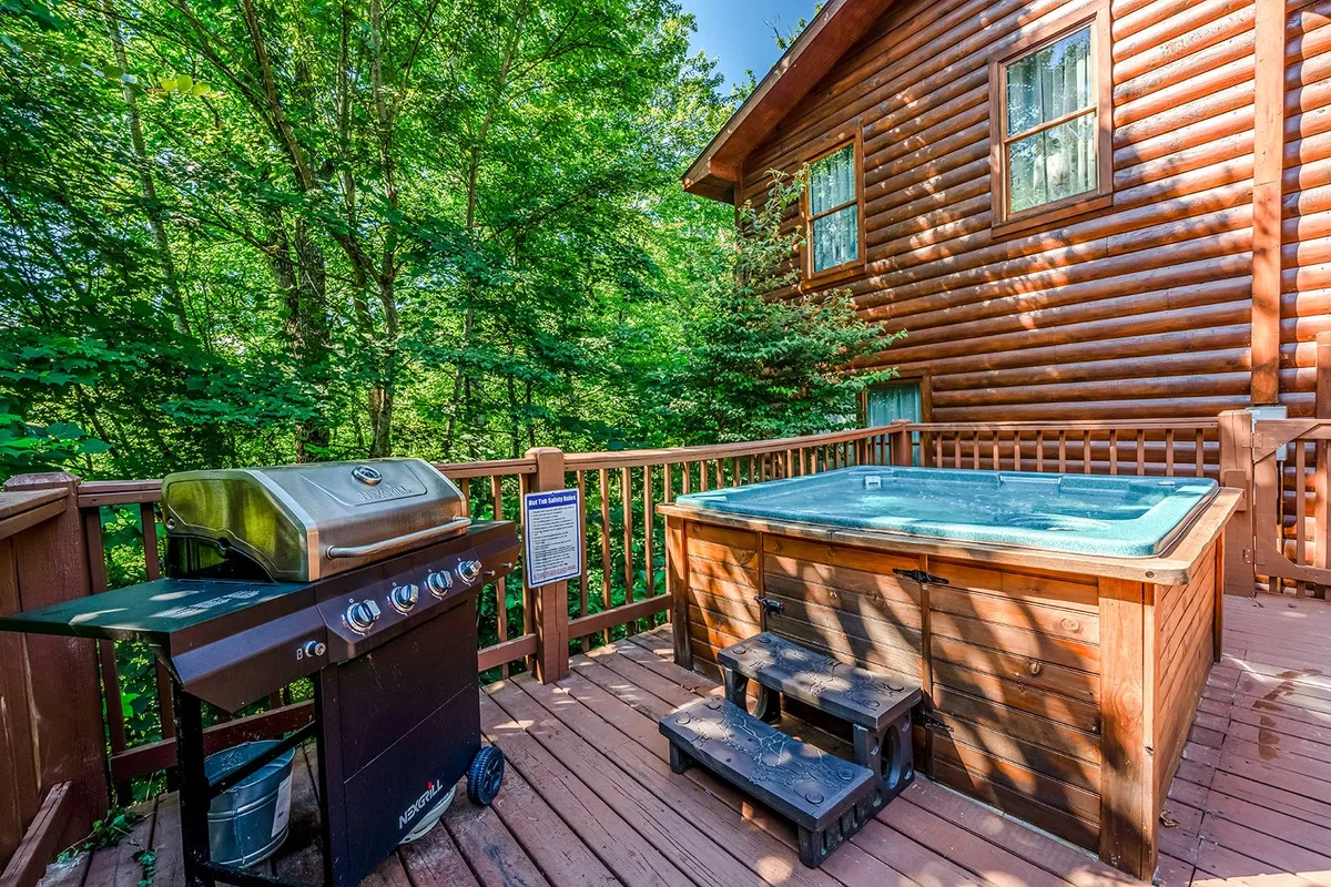 A serene exterior shot of a wooden deck with a bubbling hot tub, a grill for barbecues, and dense greenery all around. The scene suggests both relaxation and lively group meals, set against a tranquil natural backdrop.